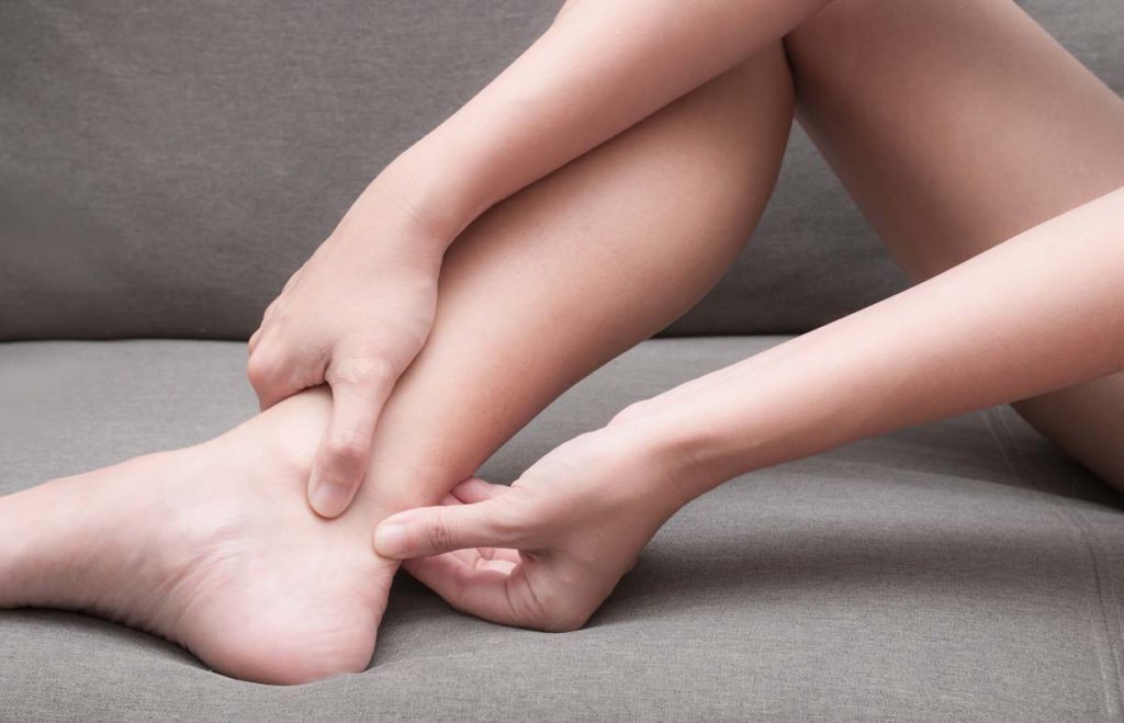 A Female Using Both Thumbs Pressing On The Back Of Her Ankle Due To The Pain Caused By Achilles Tendinitis